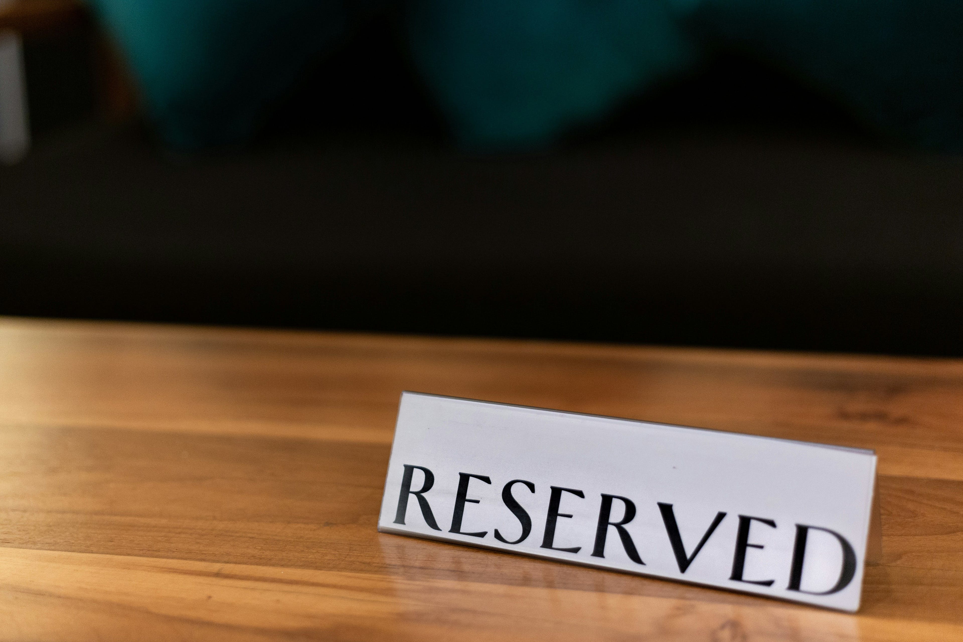 A close-up view of a 'RESERVED' sign placed on a polished wooden table in a restaurant, with plush teal cushions in the background, signaling an exclusive dining space awaiting its guests.