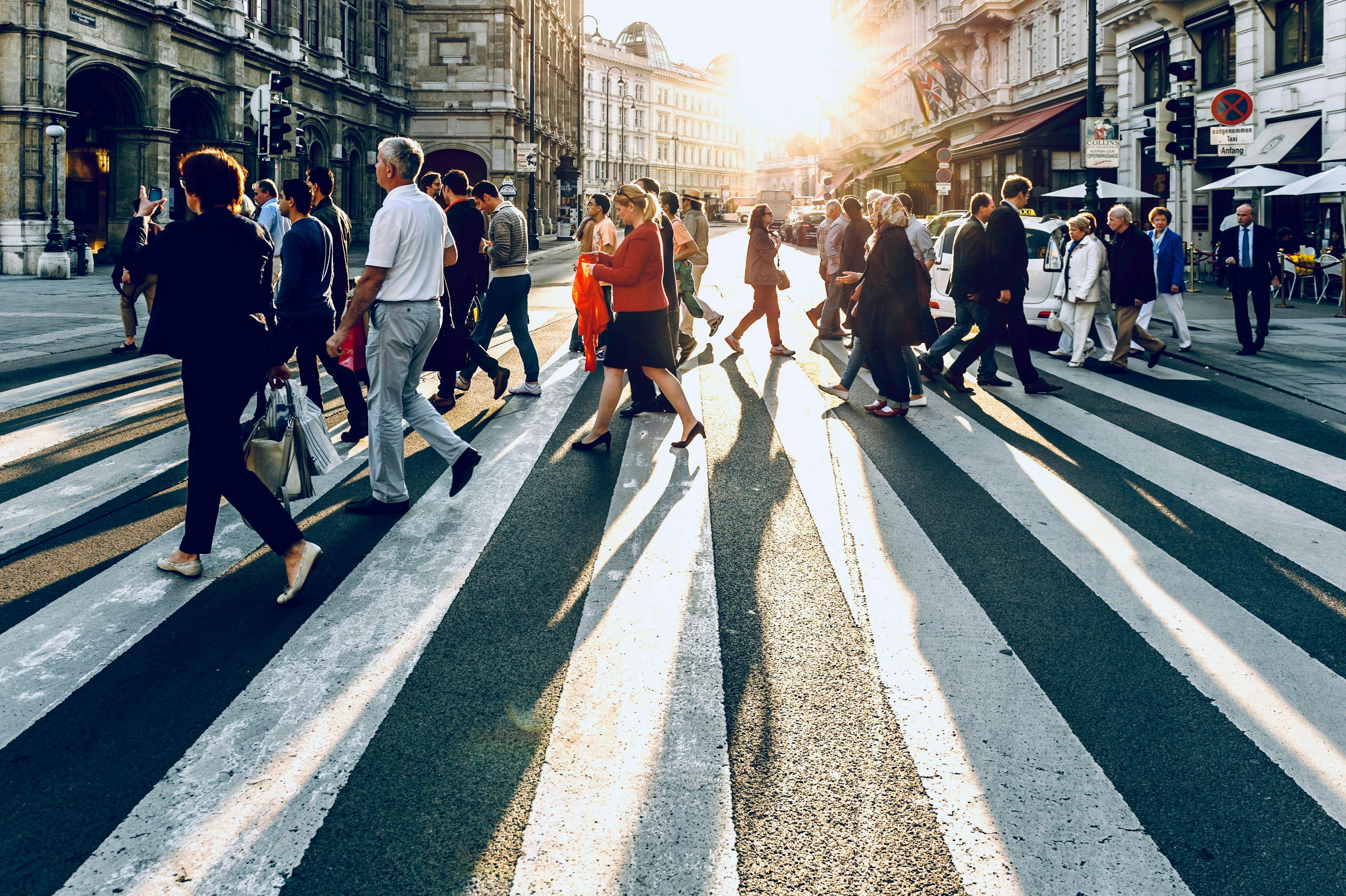 A dynamic scene of pedestrians crossing a zebra crossing in a bustling city street at sunset. The long shadows and golden sunlight create a vivid contrast with the white stripes of the crosswalk, highlighting the movement and rhythm of urban life.
