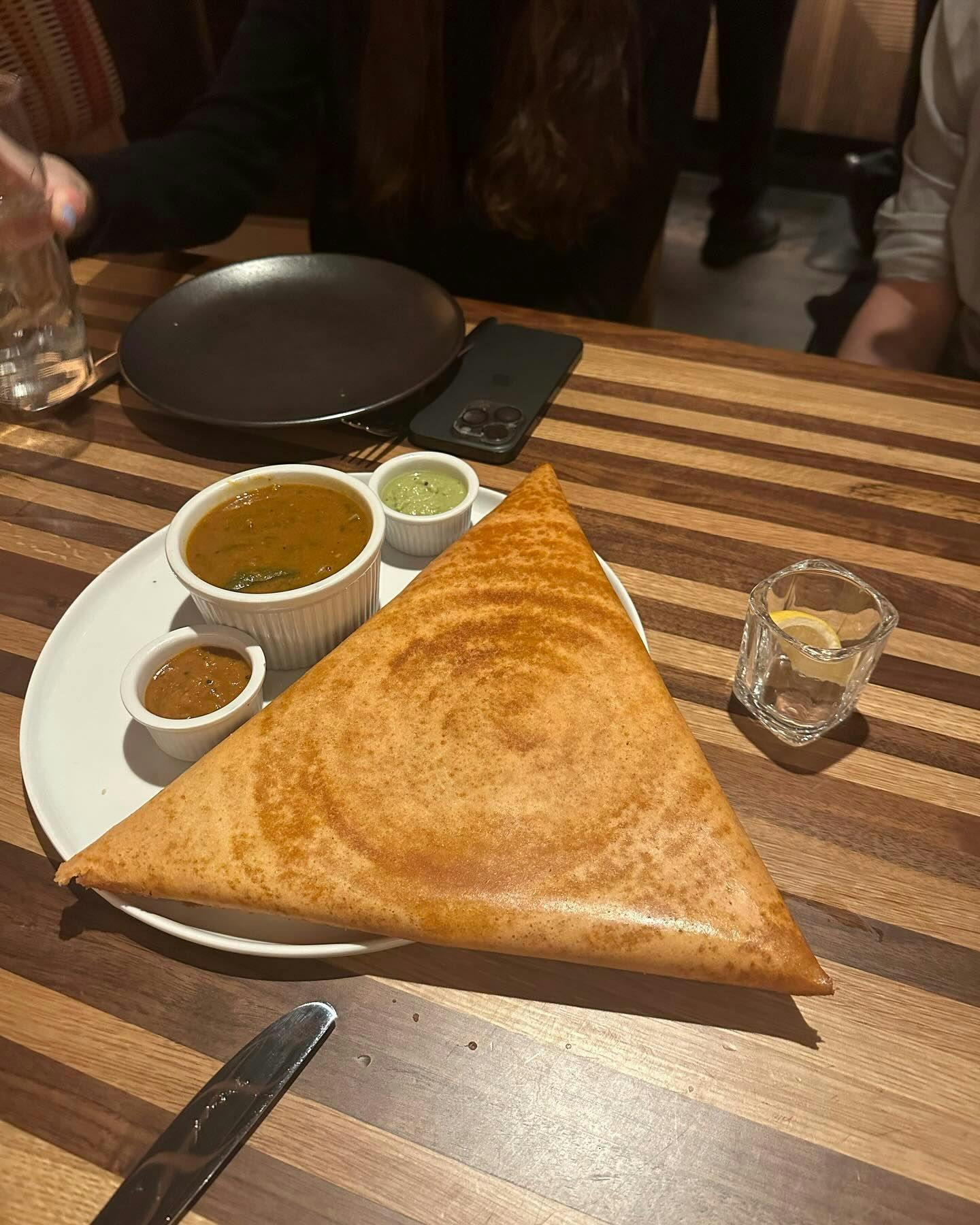 A large, golden South Indian dosa, perfectly crisp and served with a trio of chutney and sambar, presented on a classic white plate at a premium New York dining spot, available for booking on the best restaurant marketplace.