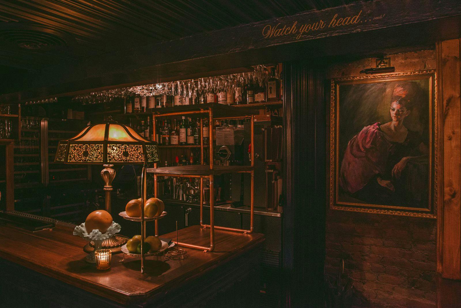 An atmospheric photograph of a cozy, dimly-lit bar interior evoking a vintage feel. Dominated by warm, muted lighting, a classic stained-glass lamp casts a soft glow on the polished wooden counter, which is adorned with a tiered stand holding fresh citrus fruits. Assorted bottles and glassware gleam subtly on the shelves in the background. The scene is complemented by a framed oil painting of a contemplative individual, adding to the rich, historical ambiance. Above the bar, the phrase 'Watch your head' is playfully inscribed, cautioning patrons of the low overhead beam.