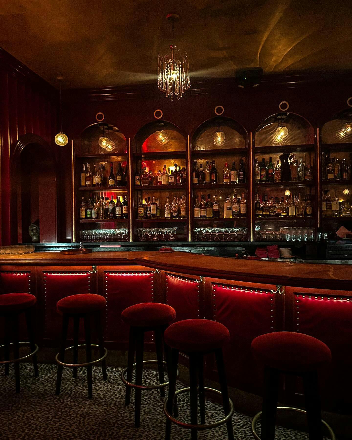 A bar and lounge bathed in a warm, inviting glow, featuring opulent chandeliers and plush red seating that echo the vintage glamour ideal for a sophisticated night out, available to reserve through Booked in New York.