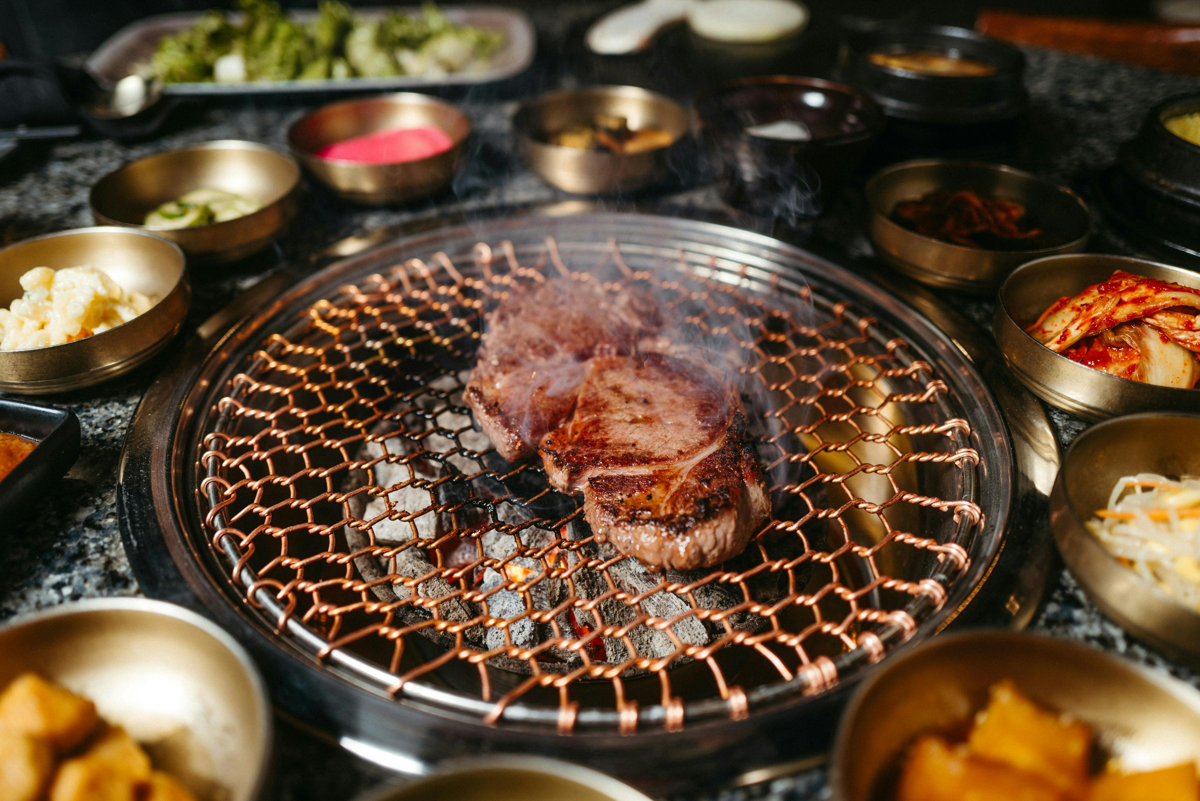 Sizzling grilled meat on a Korean BBQ with an array of traditional banchan side dishes, exemplifying the unique culinary experiences available for booking in New York through the premier reservation marketplace, Booked.